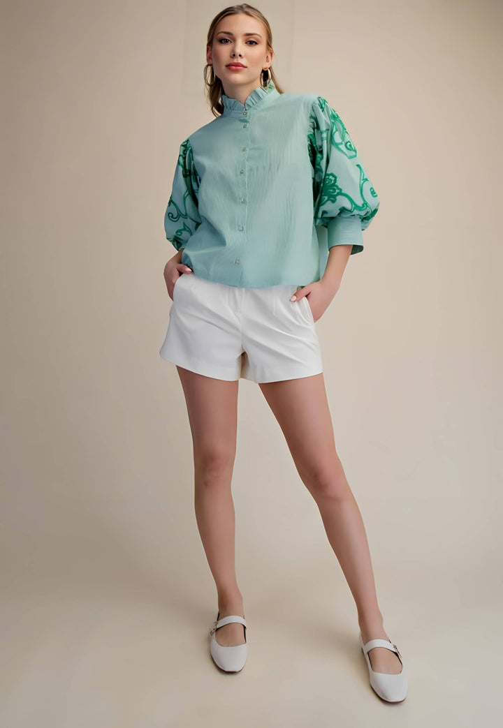 Woman wearing a light blue blouse with green patterns and white shorts on a beige background