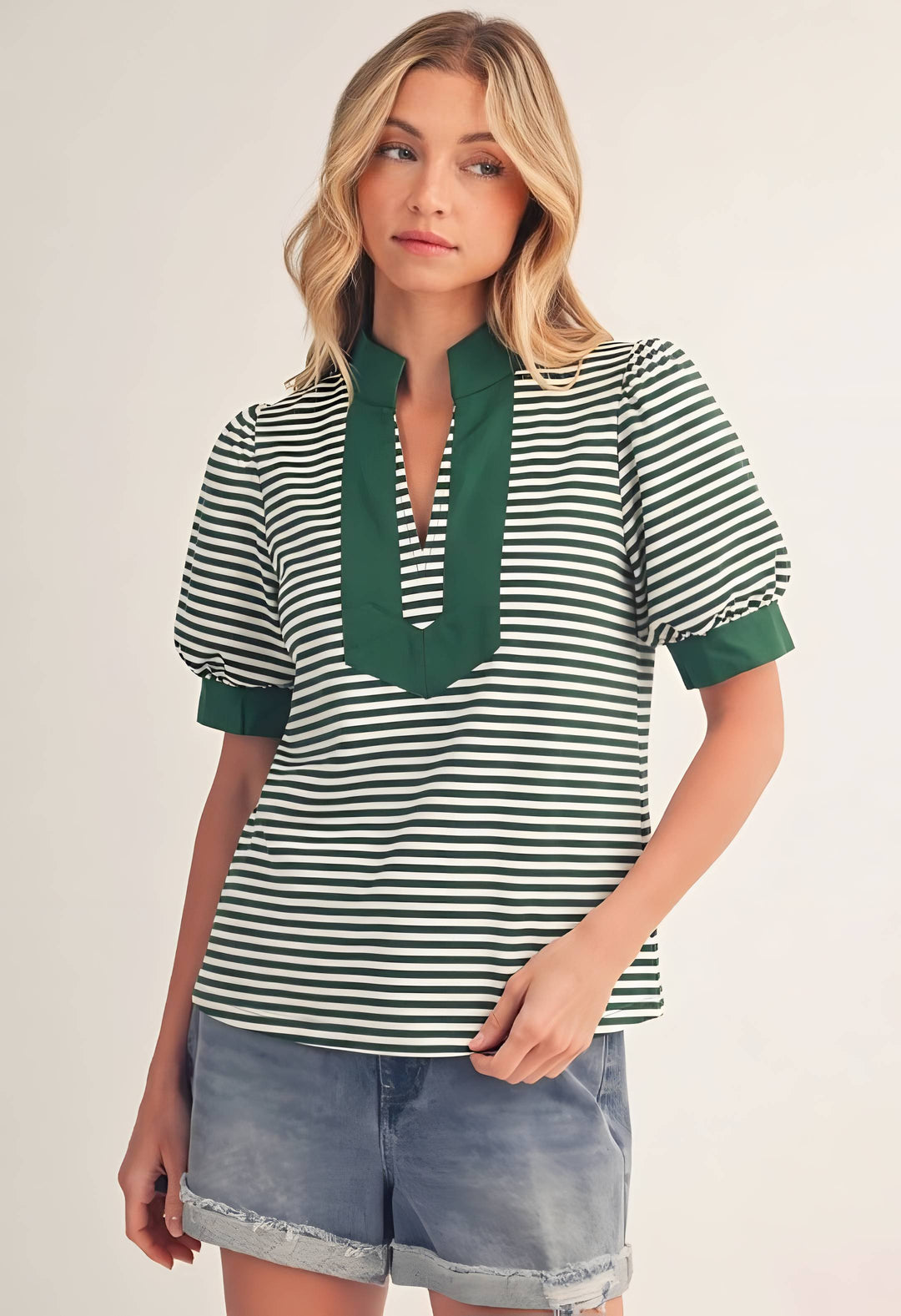 A woman modeling a short-sleeve top with green and white horizontal stripes, featuring a solid green mock neck and green rolled cuffs, paired with denim shorts against a light beige background.