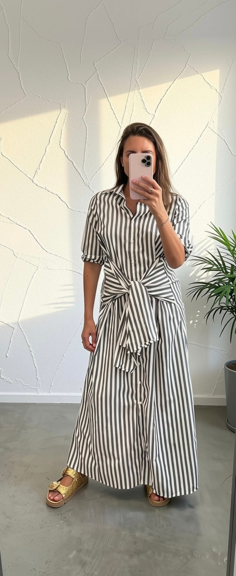 A woman takes a mirror selfie wearing a black and white vertically striped shirt dress with a tie waist, paired with gold sandals, in a modern room with a textured white wall.