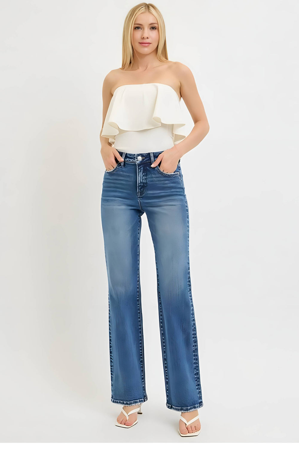 A woman modeling a pair of medium-wash blue straight-leg jeans with a ruffled strapless cream top and white heeled sandals, standing against a plain white studio background.