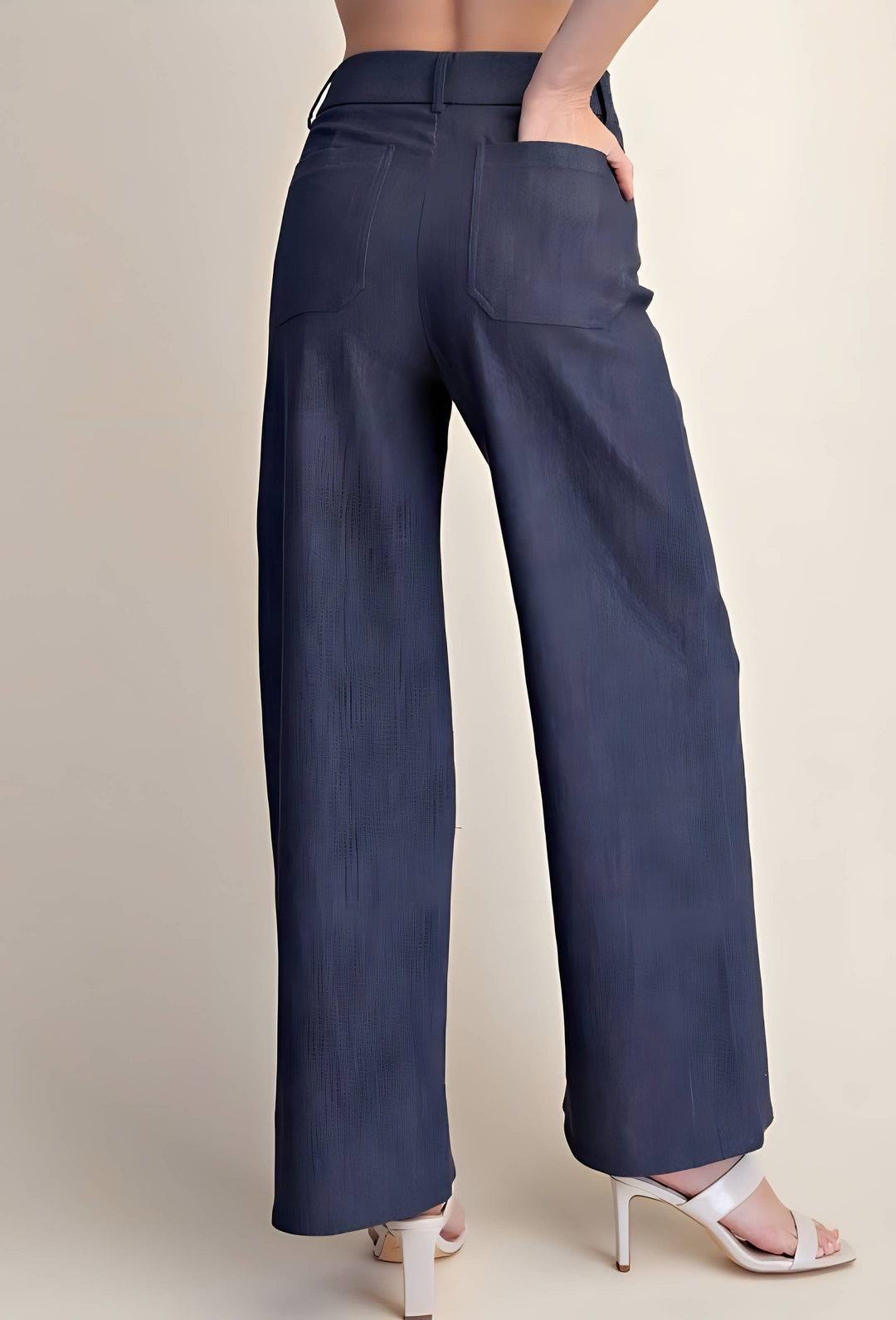 A person shown from the back wearing navy wide-leg trousers and white heeled sandals against a plain light beige studio background.