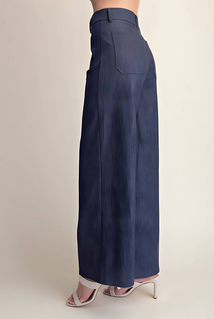 A model is shown from the waist down wearing a pair of navy blue wide-leg trousers with a pleated texture, paired with white heeled sandals, against a plain beige background.