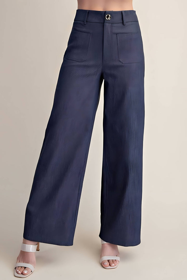 A model is wearing a pair of navy blue wide-leg trousers with a subtle vertical textured stripe pattern, paired with white heeled sandals, against a solid beige studio background.