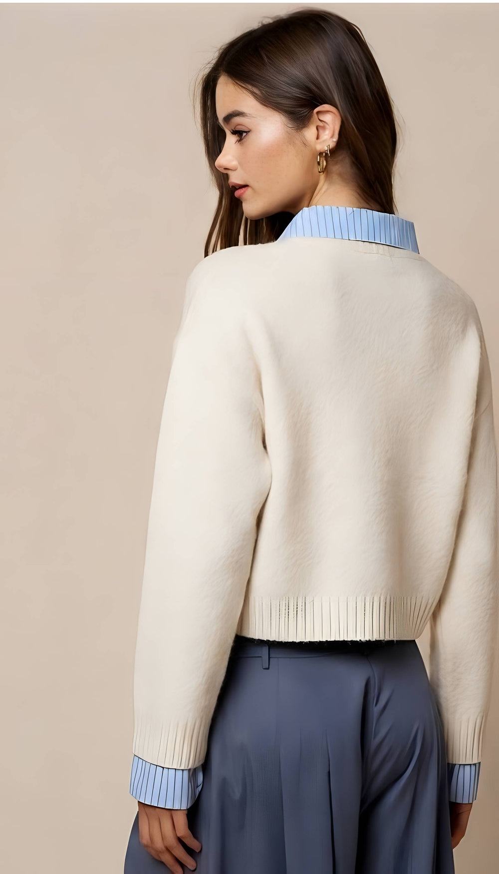 A woman shown from the back wearing a cream knit cropped sweater with light blue striped collar and cuffs, paired with slate blue pants, against a solid beige background.
