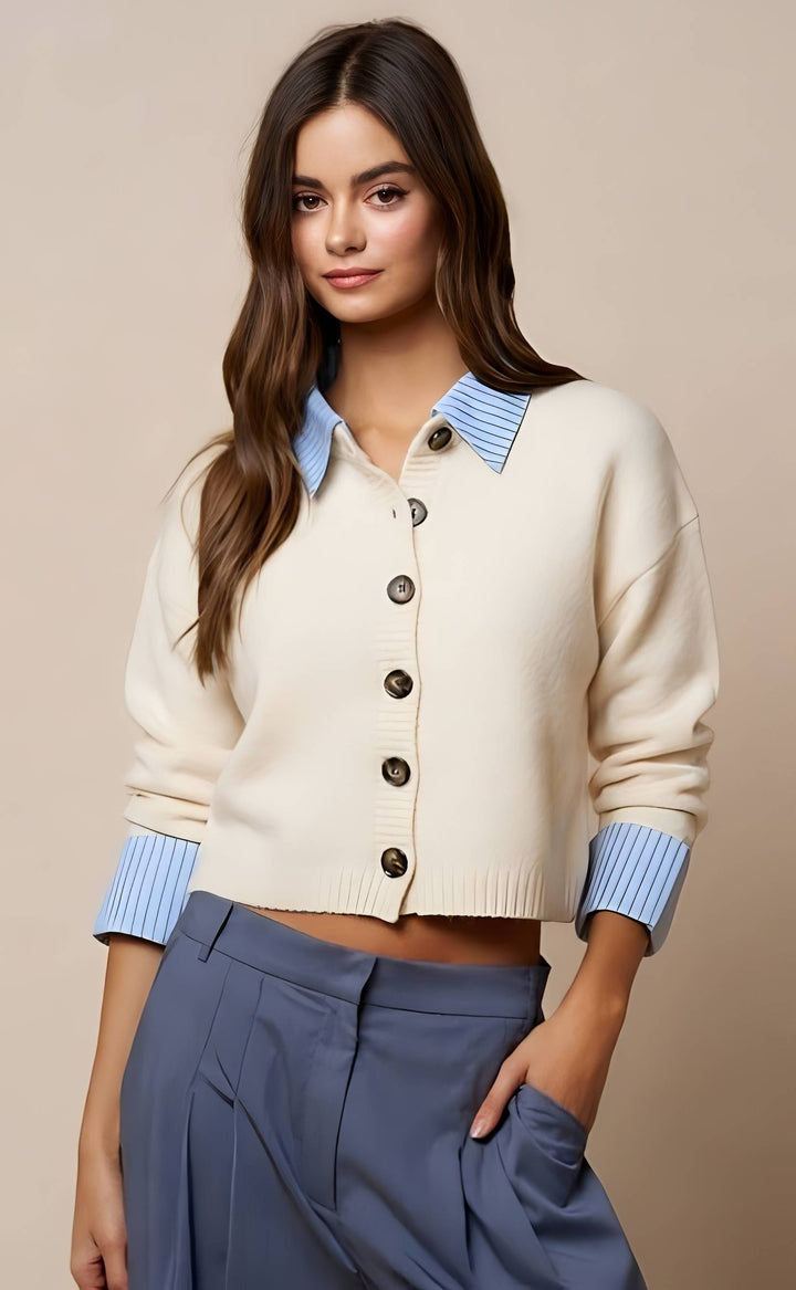 A woman modeling a cropped cream cardigan with light blue striped collar and cuffs, paired with slate blue pants, against a solid beige background.