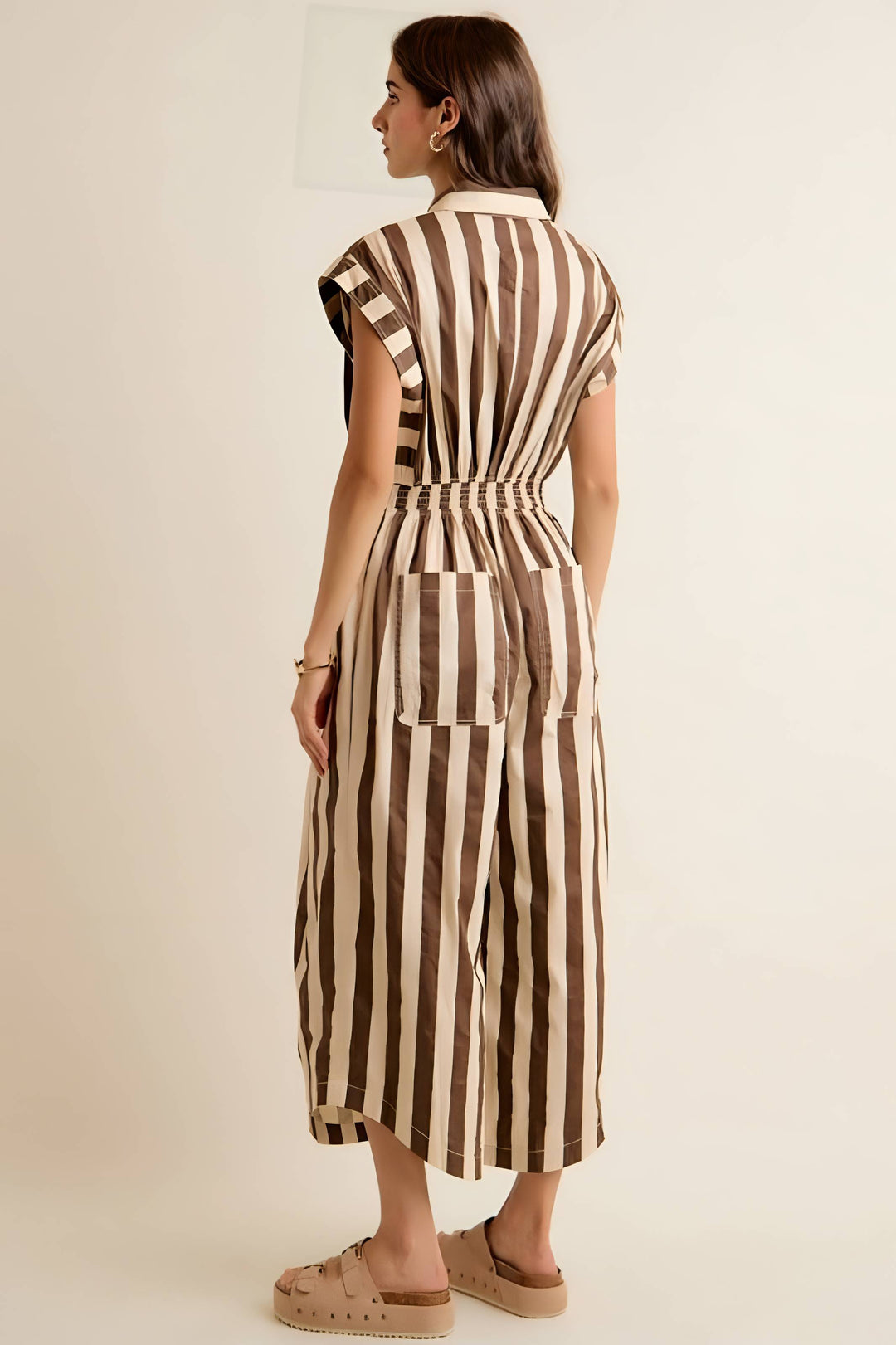 A woman is shown from the back wearing a brown and beige vertically striped midi dress with a tie waist and sleeveless cap sleeves, paired with beige platform sandals, against a plain cream background.