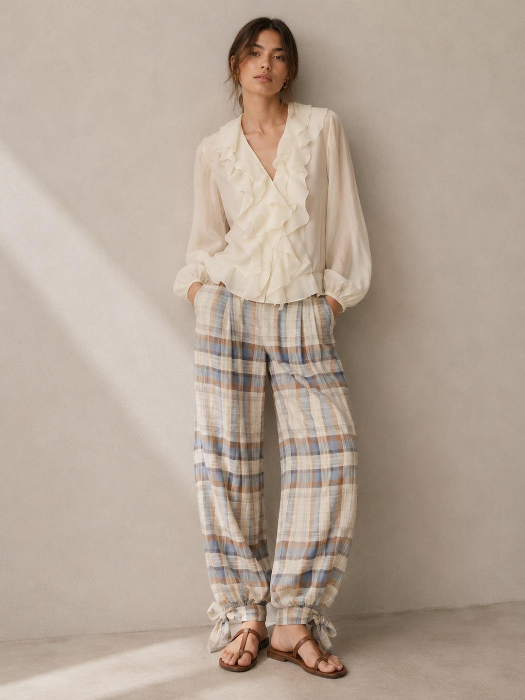 A woman stands against a textured beige wall wearing a cream ruffled blouse and beige plaid trousers with blue and brown lines, paired with brown leather sandals.