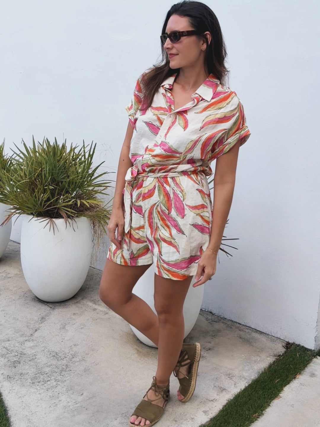 Woman in a colorful romper standing outdoors with a white wall and plant in the background