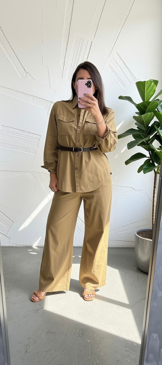 A woman takes a mirror selfie wearing a tan two-piece set consisting of a long-sleeve button-up shirt with a thin black belt and matching wide-leg pants, standing in a bright room with a potted plant.