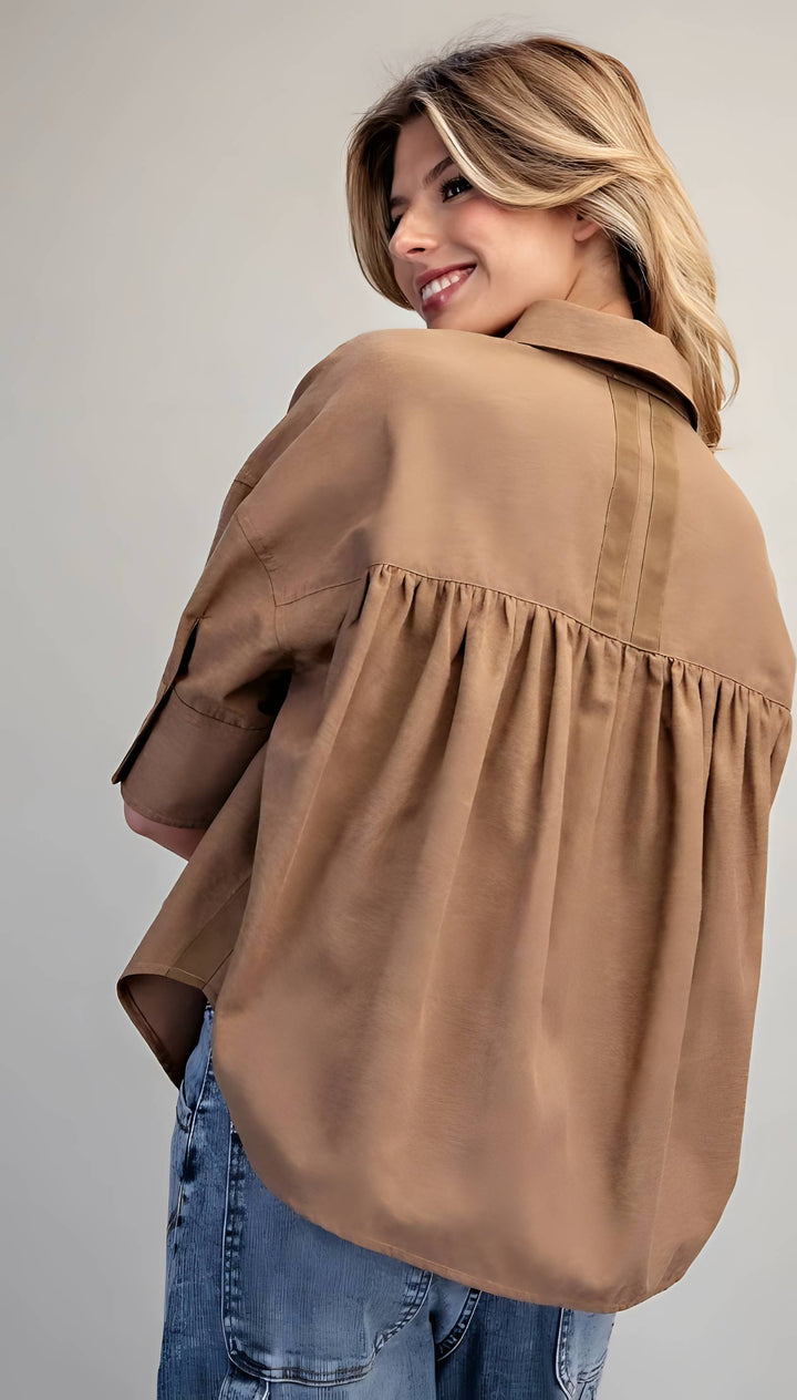 A woman wearing an oversized tan button-up blouse with a gathered waist seam and blue jeans, photographed against a plain light gray studio background.