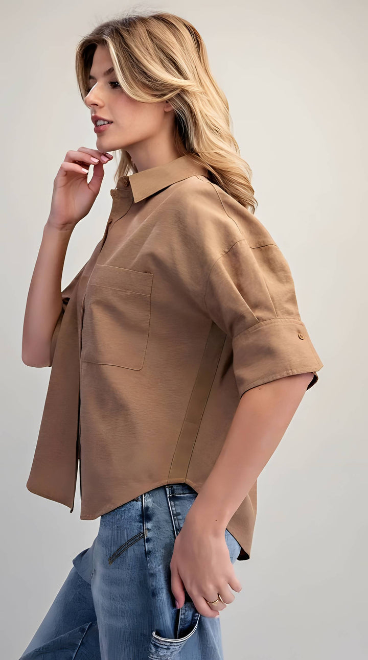 A woman wearing a tan short-sleeve button-up shirt with blue jeans, photographed against a plain light gray studio background.