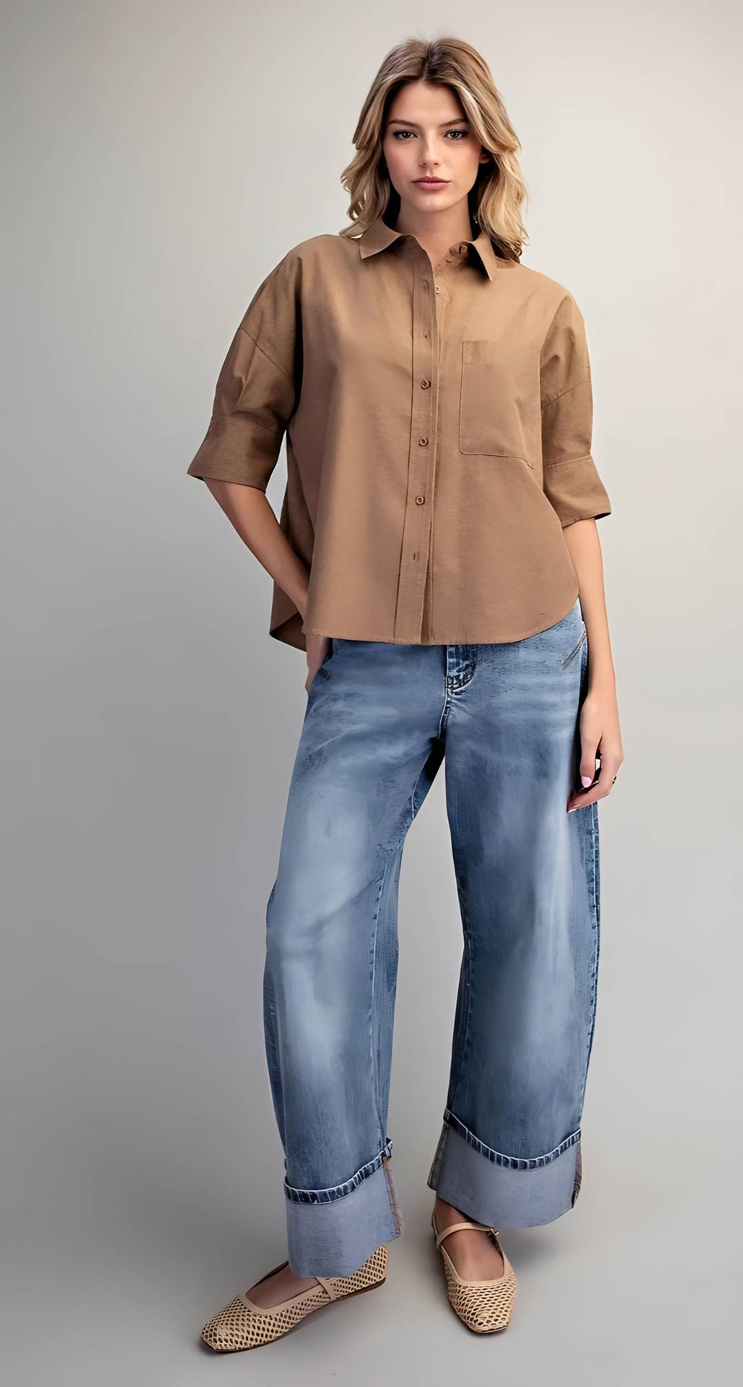 A woman modeling a tan short-sleeve button-up shirt with light blue wide-leg jeans and woven flat shoes, standing against a neutral gray studio background.