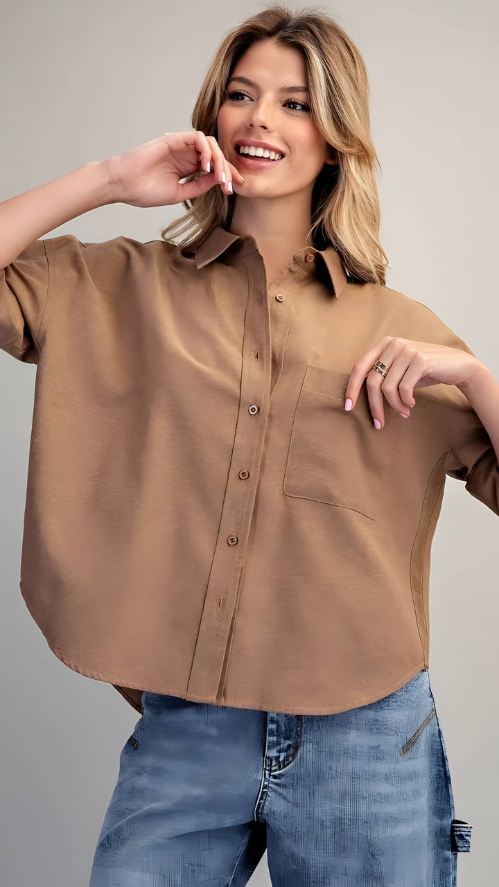A woman wearing a cropped, short-sleeve button-up shirt in a warm camel brown color, paired with blue jeans, against a solid light gray background.