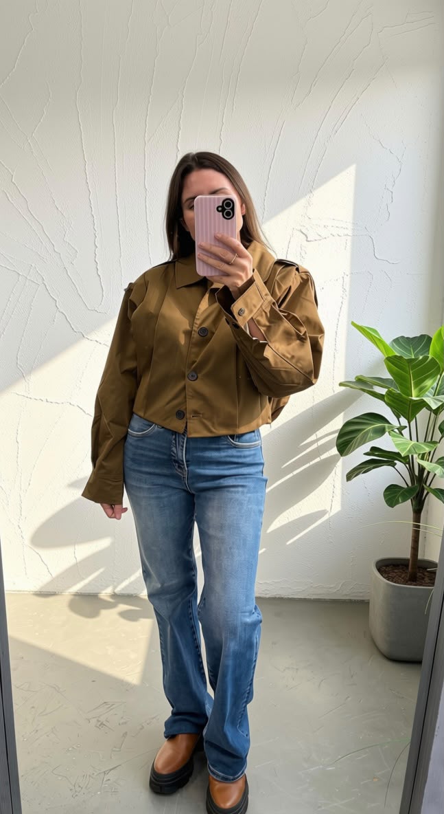 A woman takes a mirror selfie wearing a cropped brown button-up jacket with puff sleeves and blue jeans, standing in a brightly lit room with a potted plant in the background.