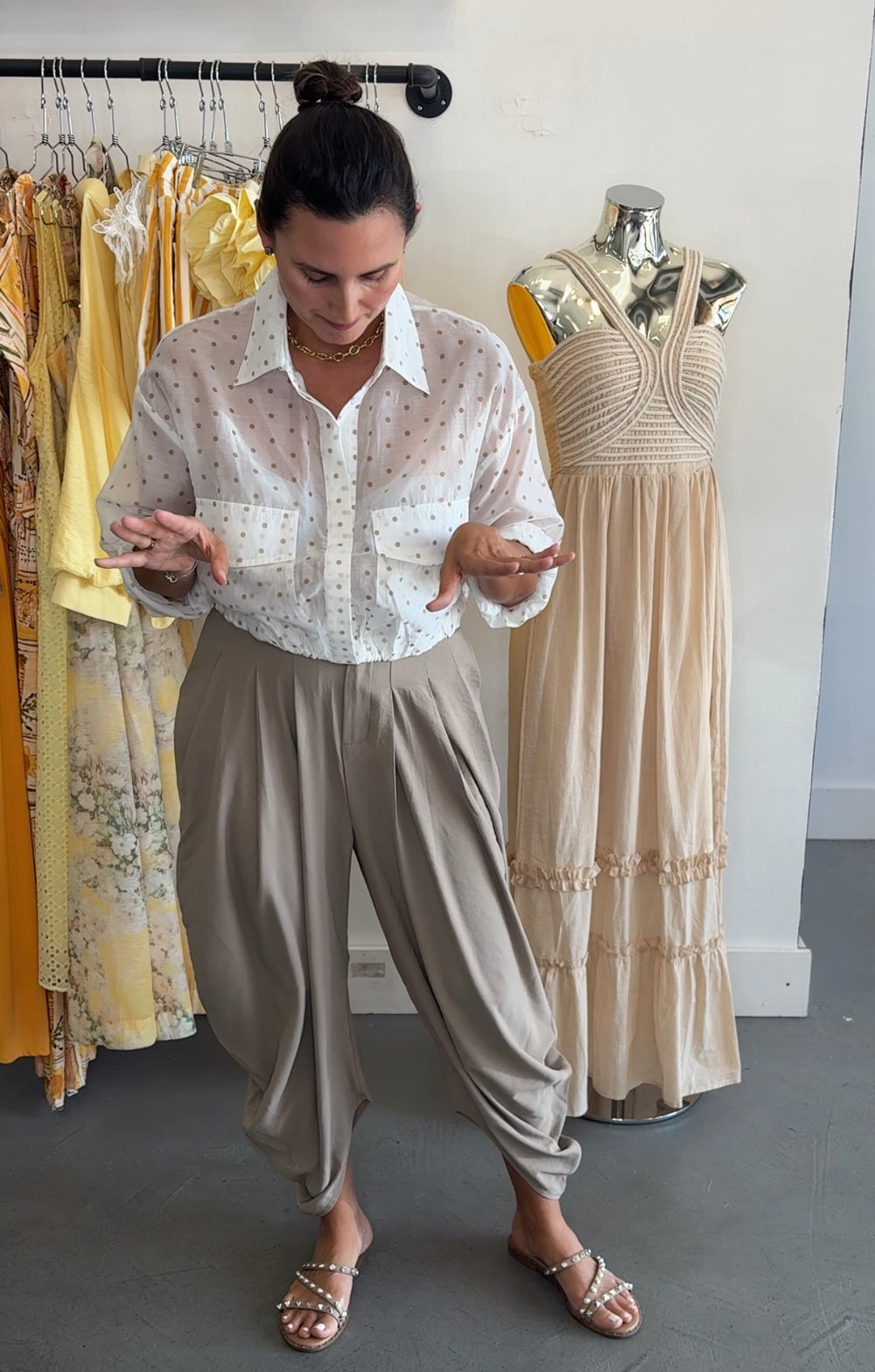 A woman modeling a white sheer long-sleeve button-up shirt with tiny red polka dots, paired with loose taupe-brown harem pants and metallic sandals, standing in a boutique next to a mannequin wearing a beige dress.
