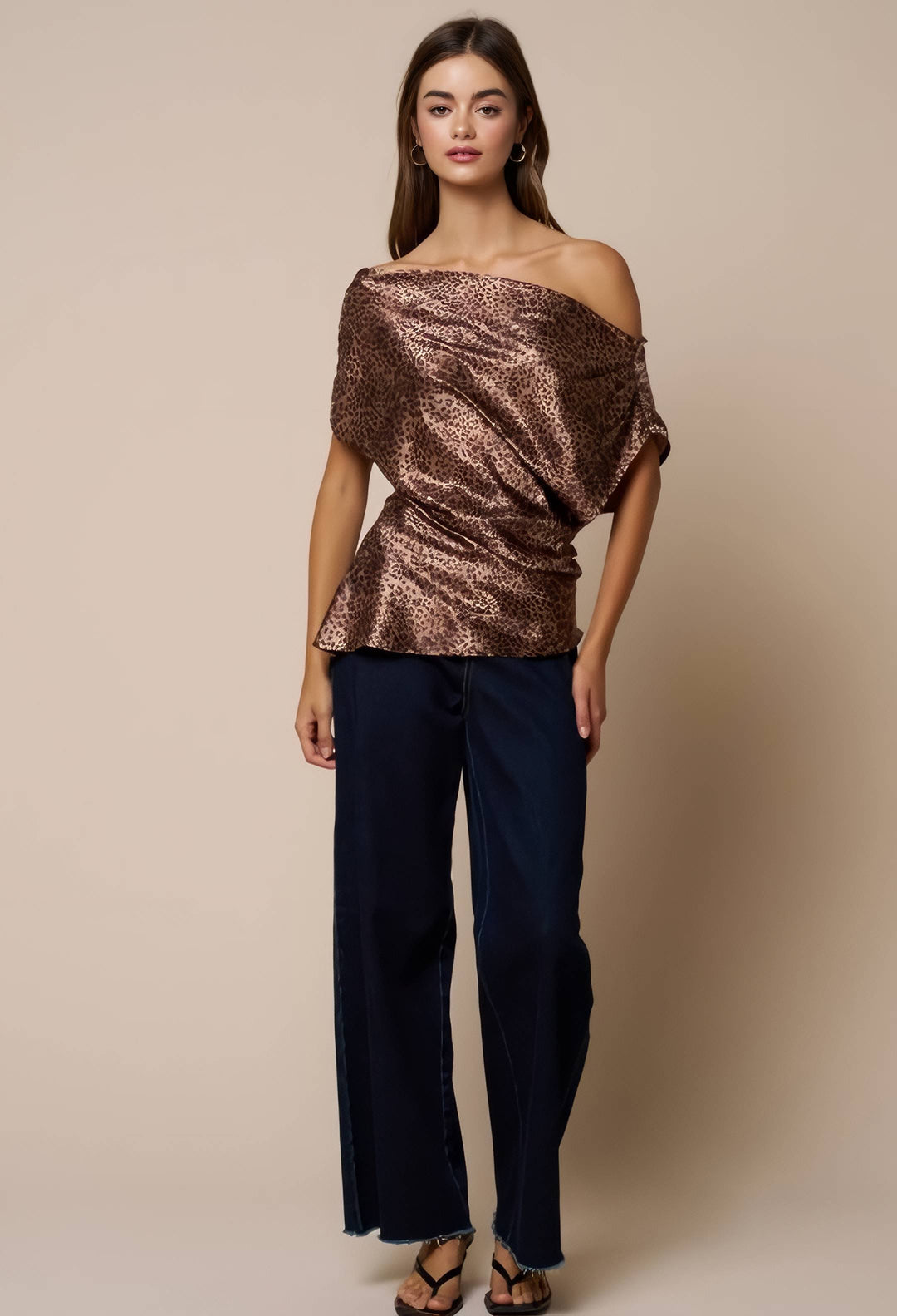 A woman modeling an off-the-shoulder metallic bronze sequined top paired with dark navy wide-leg pants and brown strappy sandals, set against a solid beige studio background.