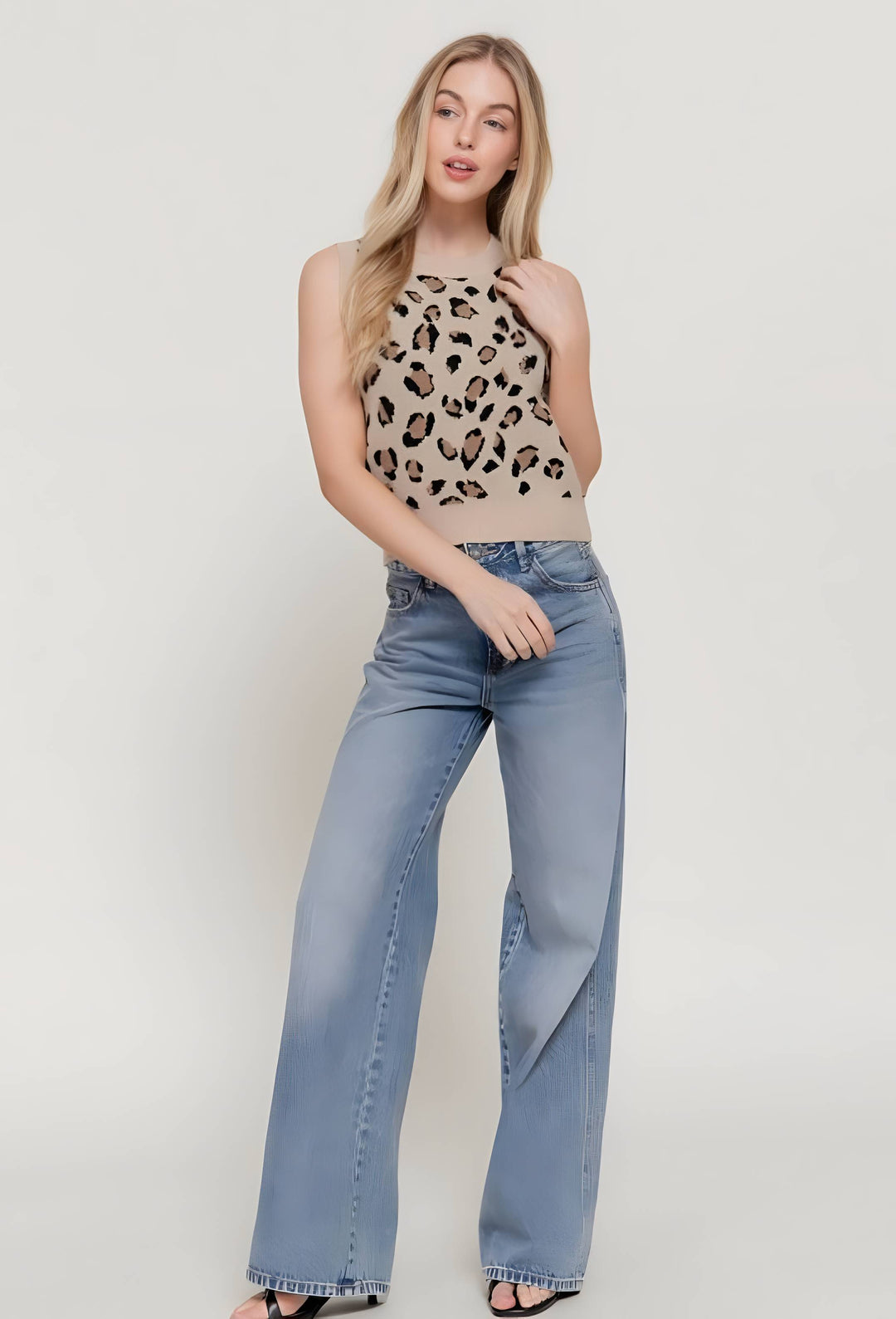 A woman modeling a sleeveless beige knit top with a black leopard print pattern and light-wash wide-leg blue jeans against a plain light gray studio background.