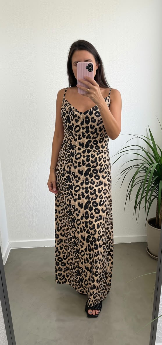 A woman takes a mirror selfie wearing a sleeveless leopard-print maxi dress with thin straps and black heeled sandals in a minimalist room with a potted plant.