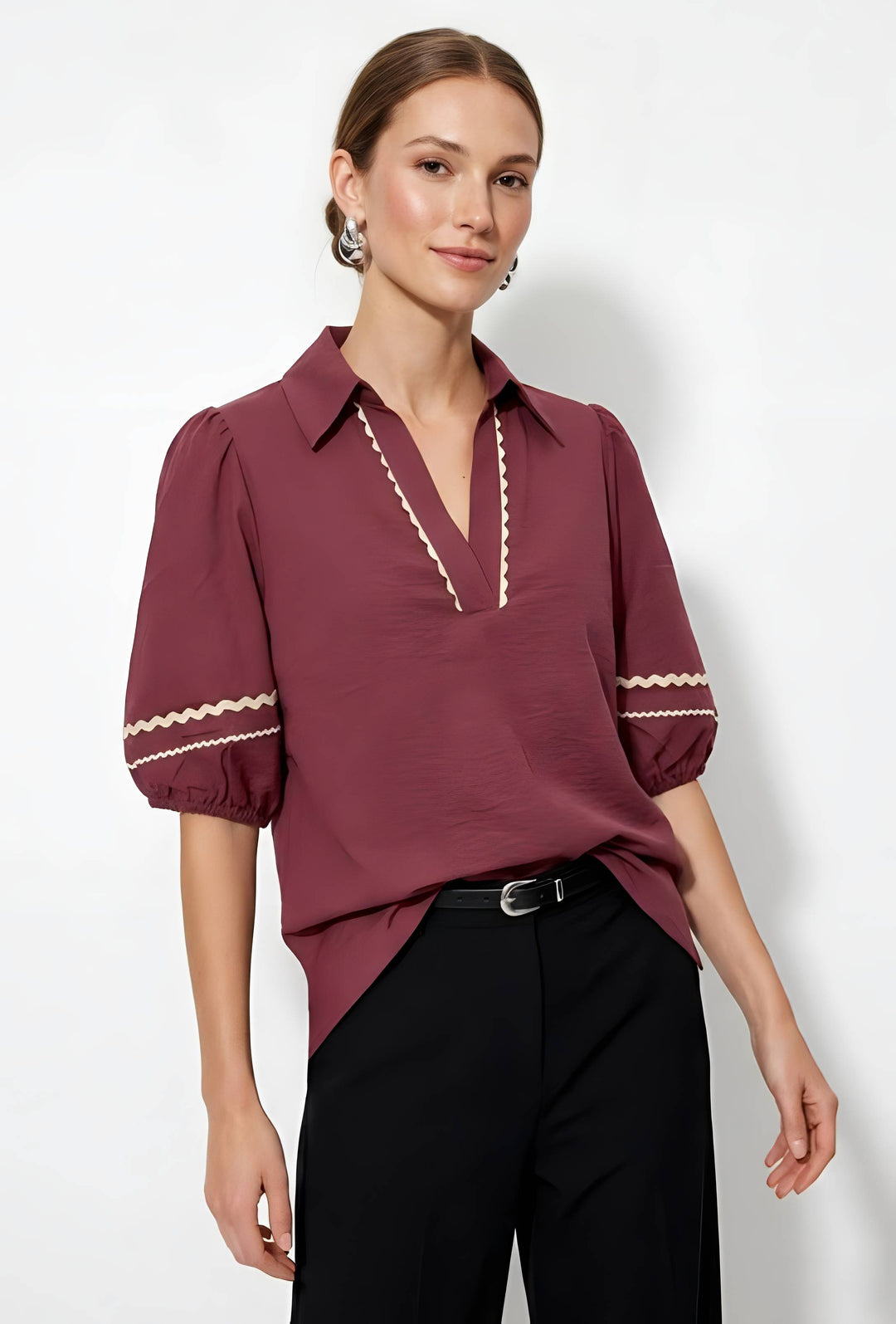 A woman modeling a burgundy short-sleeve blouse with cream zigzag trim details on the collar placket and sleeve cuffs, paired with black trousers, against a plain white studio background.