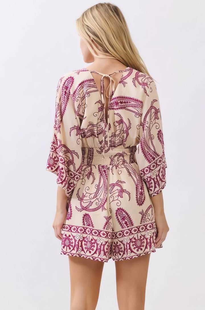 A woman shown from the back wearing a short romper with a cream base and a magenta paisley pattern, featuring a tie-back neckline and ruffled sleeves.