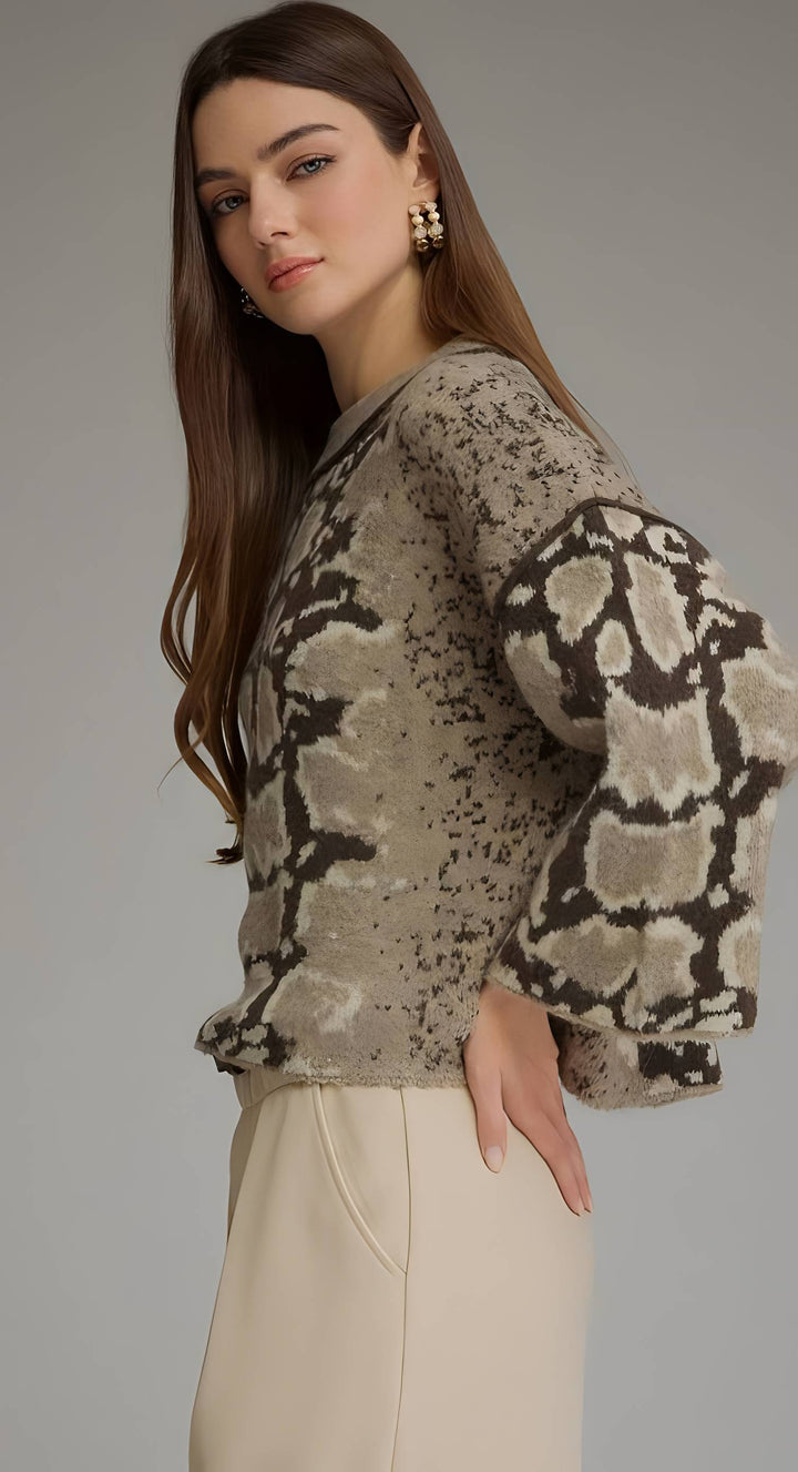 A woman modeling a cropped knit sweater with a beige, brown, and black animal print pattern, paired with cream-colored trousers, against a solid gray studio background.