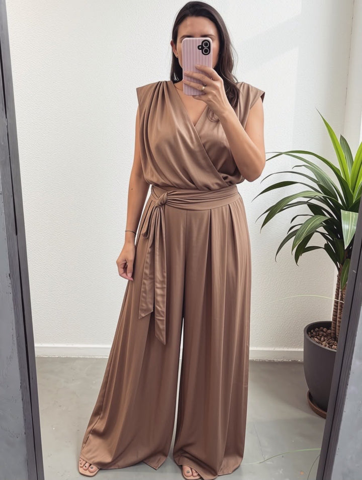 A woman takes a mirror selfie wearing a sleeveless, wrap-style pant set in a warm taupe-brown satin-like fabric with a tied waist belt and wide legs. The setting is indoors with a white wall and a potted plant visible on the right.