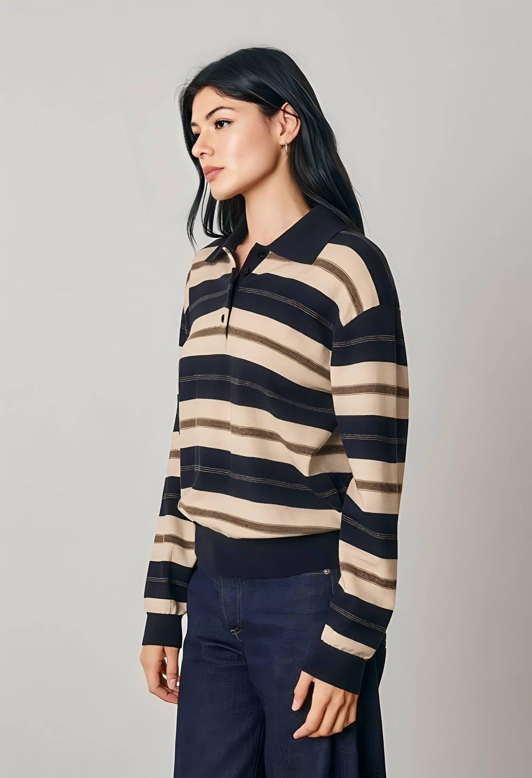 A woman wearing a navy and beige horizontally striped long-sleeve knit polo sweater, paired with dark blue trousers, photographed against a neutral light gray studio background.