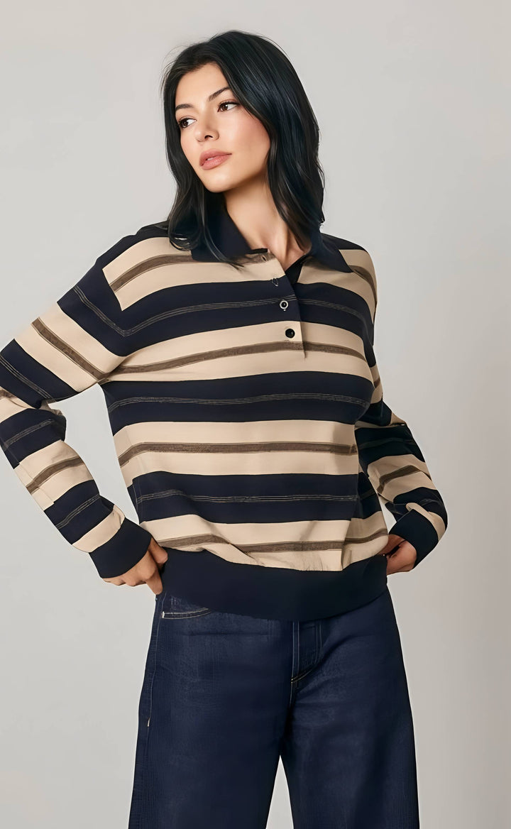 A woman modeling a long-sleeve knit polo sweater with horizontal stripes in beige, brown, and black, paired with dark blue jeans, against a plain light gray studio background.