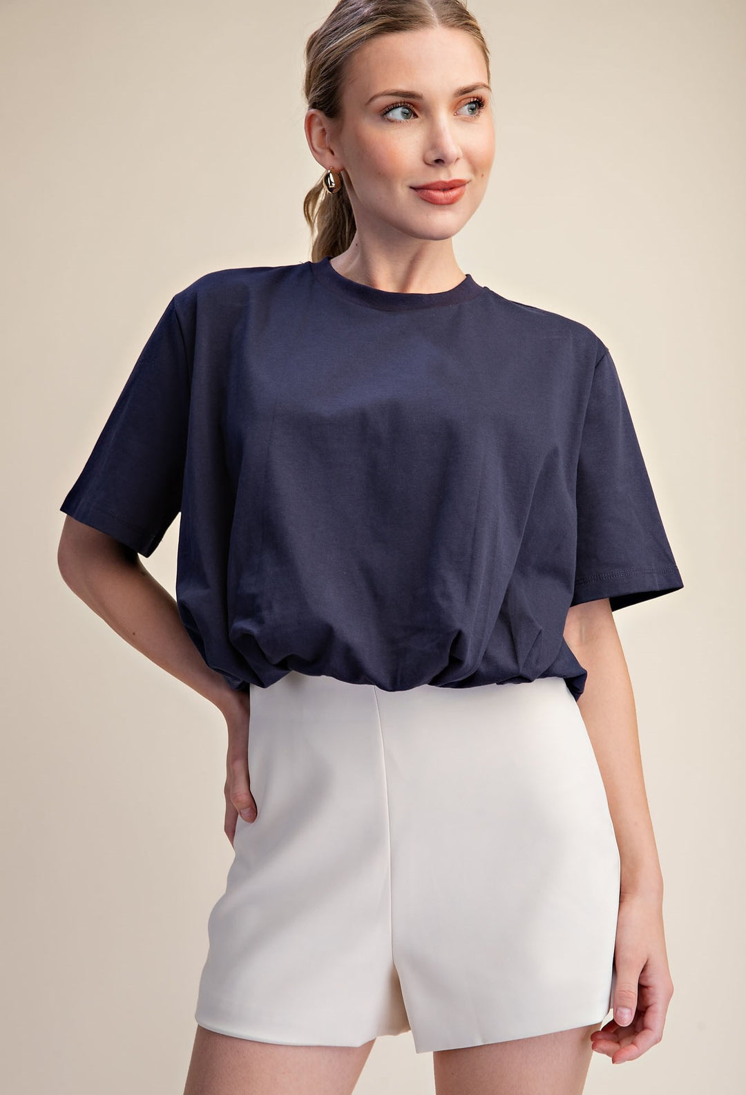 A woman wearing a navy blue short-sleeve crewneck t-shirt and off-white shorts, posing against a solid beige studio background.