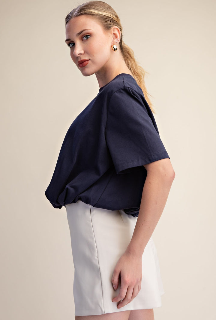 A woman modeling a navy blue short-sleeve blouse with voluminous draped sleeves and a gathered waist, paired with a white mini skirt, against a plain beige studio background.