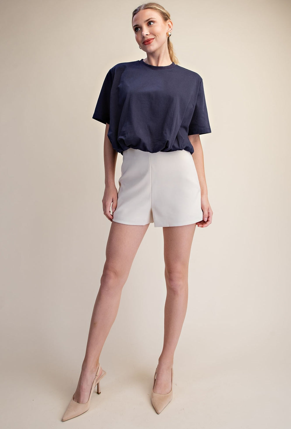A woman modeling a navy blue short-sleeve top and white shorts against a plain beige studio background.