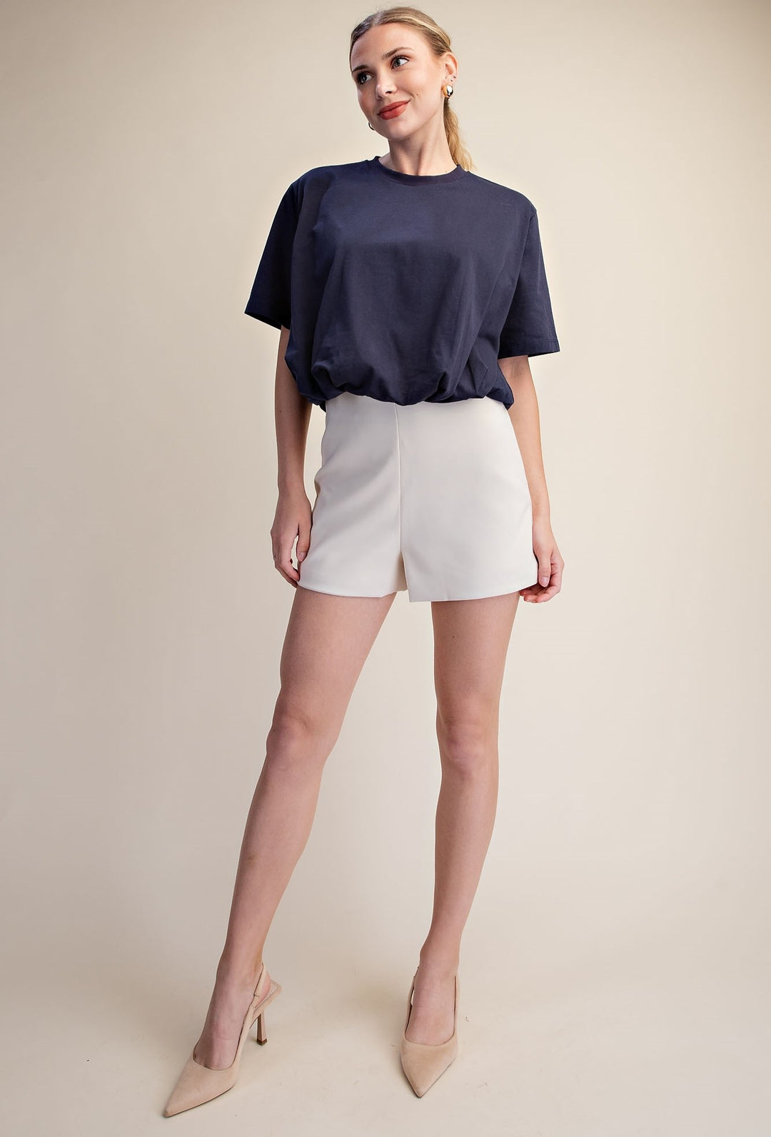 A woman modeling a navy blue short-sleeve top and white shorts against a plain beige studio background.