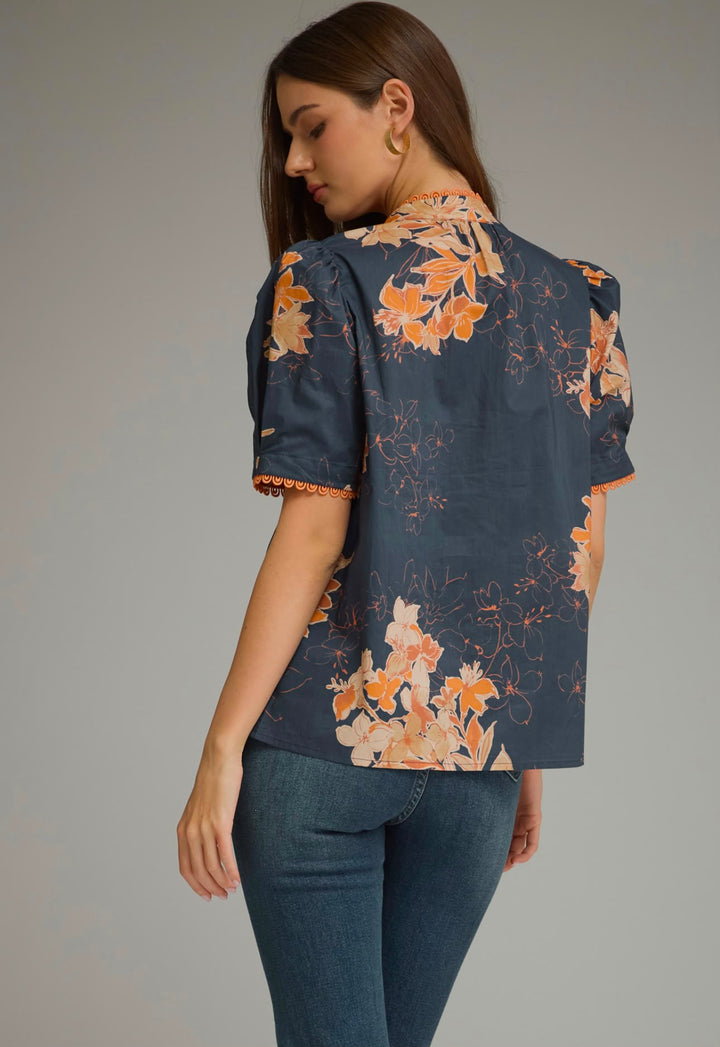 A woman shown from the back wearing a navy blue short-sleeve blouse with a peach and orange floral print, paired with blue jeans, against a solid gray studio background.