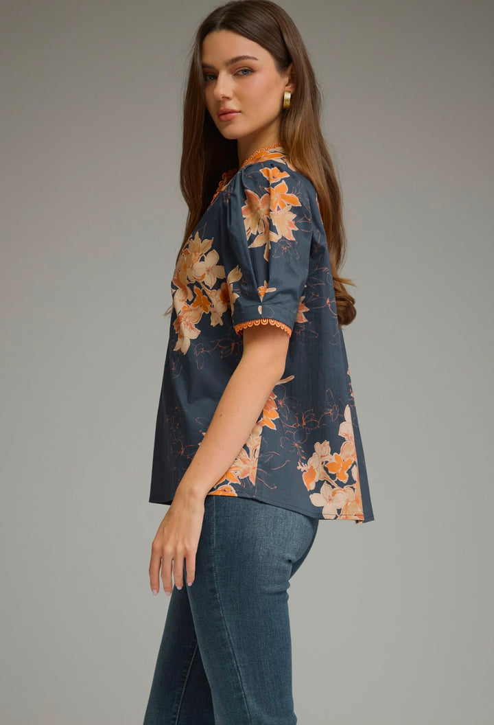 A woman wearing a short-sleeve navy blue blouse with a warm orange and beige floral print, paired with blue jeans, photographed against a neutral gray studio background.