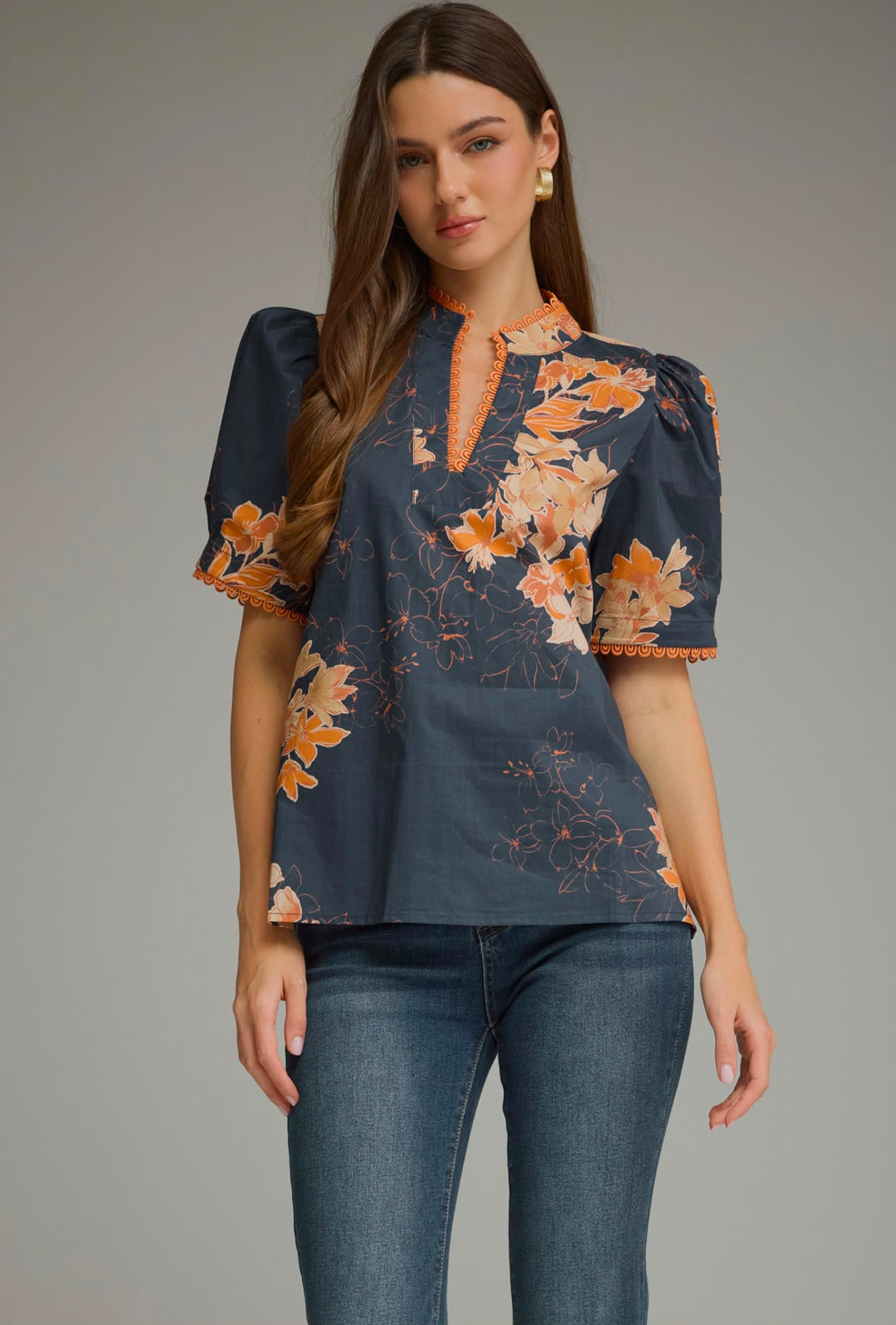 A woman modeling a short-sleeve blouse with puff sleeves, featuring a dark slate blue base with orange and peach floral patterns and orange trim, paired with blue jeans against a neutral gray studio background.