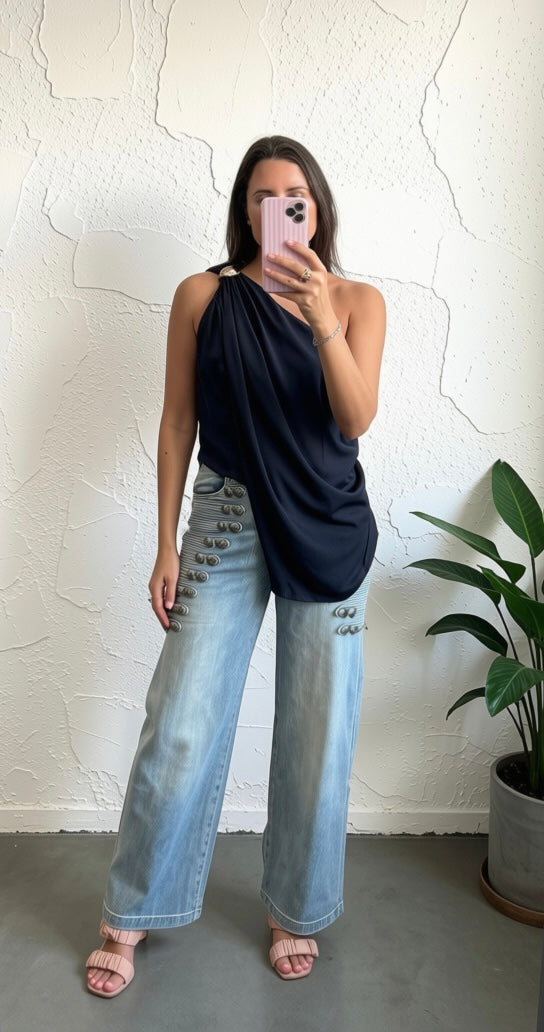 A woman takes a mirror selfie wearing a dark navy draped sleeveless top and light-wash wide-leg jeans with distressed details, standing against a textured white wall next to a potted plant.