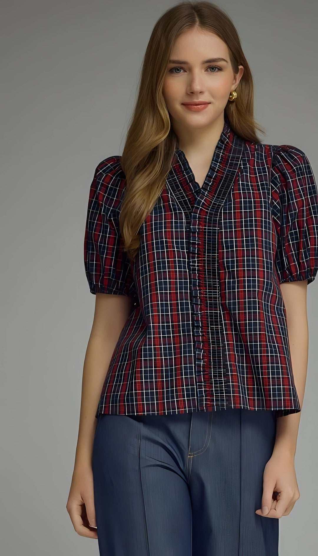 A woman modeling a short-sleeve plaid button-up shirt with puff sleeves and dark blue wide-leg trousers, photographed against a neutral gray studio background.