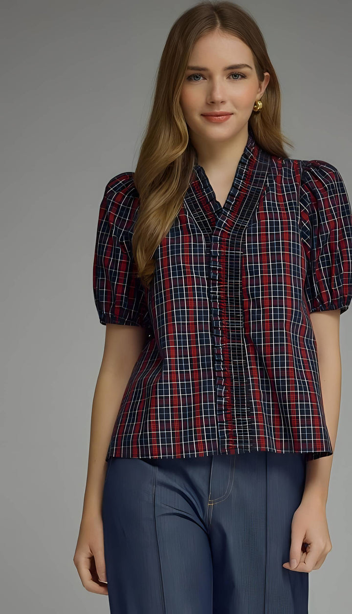 A woman modeling a short-sleeve plaid button-up shirt with puff sleeves and dark blue wide-leg trousers, photographed against a neutral gray studio background.