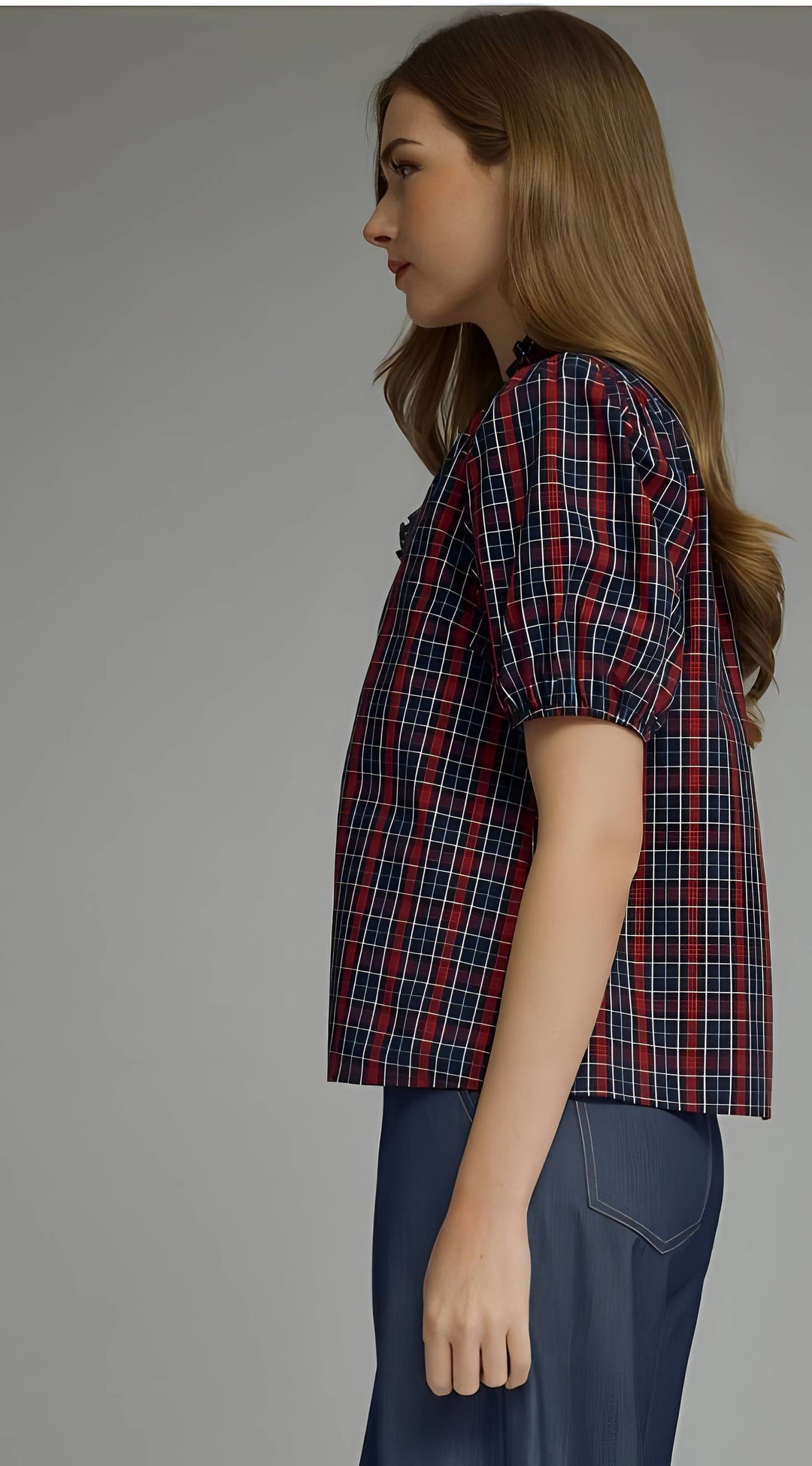 A woman shown in side profile wearing a short-sleeve plaid button-up shirt with puff sleeves in red, navy, and white, paired with navy pants, against a solid gray studio background.