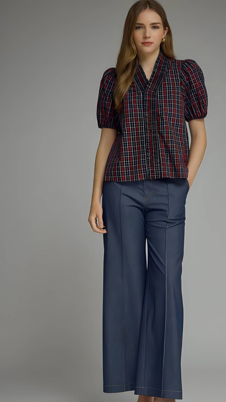 A woman modeling a short-sleeve plaid button-up shirt with puff sleeves and dark blue wide-leg trousers, photographed against a neutral gray studio background.