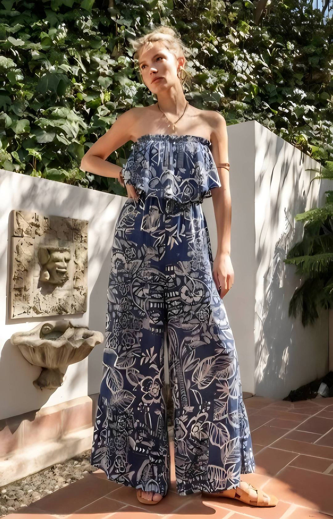 Woman wearing a blue floral strapless jumpsuit standing outdoors with greenery in the background.