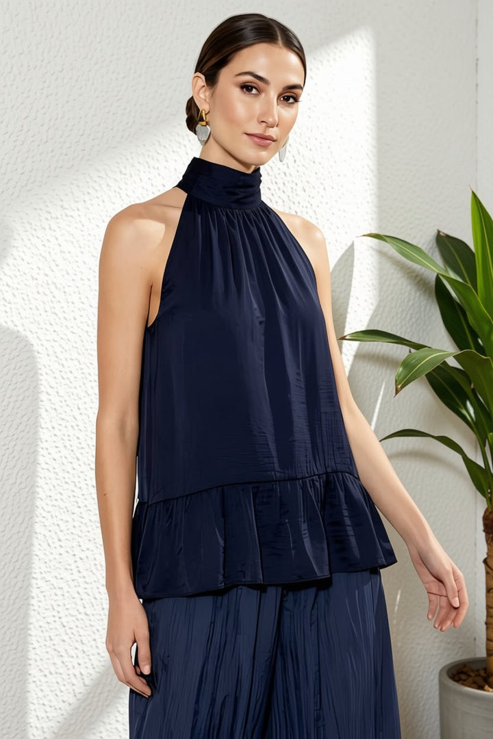 A woman modeling a sleeveless navy blue halter top with a gathered peplum waist, paired with matching pleated navy bottoms, set against a textured white wall with a green plant in the background.