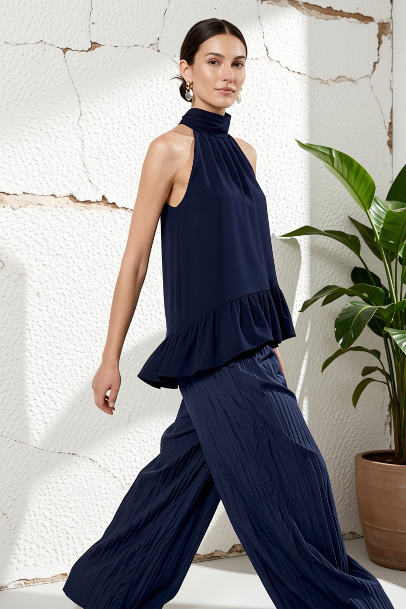 A woman models a navy blue two-piece outfit consisting of a sleeveless high-neck ruffled top and matching pleated wide-leg pants, set against a textured white wall with a potted plant.