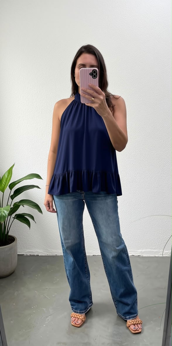 A woman takes a mirror selfie wearing a navy blue halter top with a ruffled hem, paired with blue jeans and pink sandals, in a minimalist room with a potted plant.