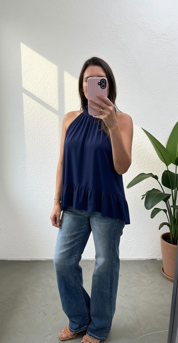 A woman takes a mirror selfie wearing a navy blue halter top with a ruffled hem and blue jeans in a bright room with a potted plant.