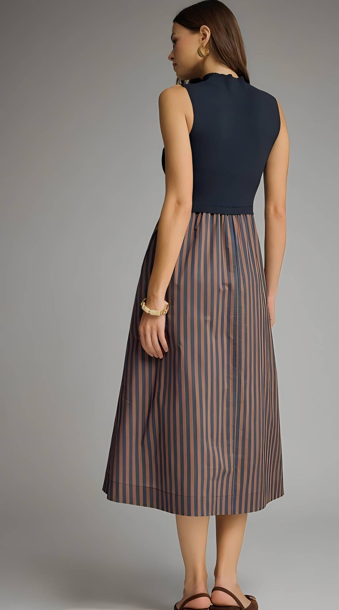 A woman shown from the back wearing a sleeveless midi dress with a solid navy top and a pleated skirt featuring vertical brown and beige stripes, set against a plain gray studio background.