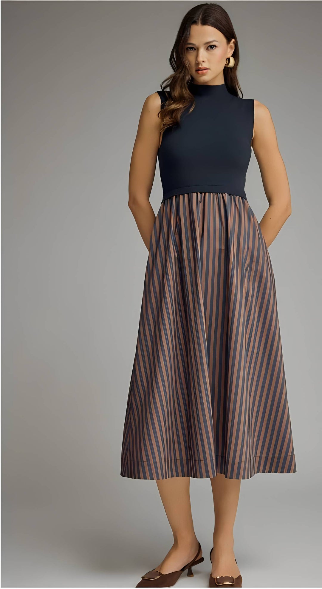 A woman modeling a sleeveless midi dress with a dark navy mock neck bodice and a pleated skirt featuring vertical stripes in brown, beige, and muted blue, paired with brown pointed-toe flats against a neutral gray studio background.