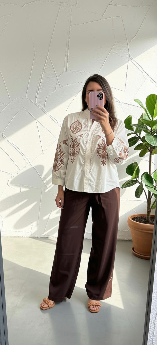 A woman takes a mirror selfie wearing a cream-colored embroidered long-sleeve blouse with brown floral patterns and dark brown wide-leg pants, standing in a brightly lit room with a potted plant in the background.