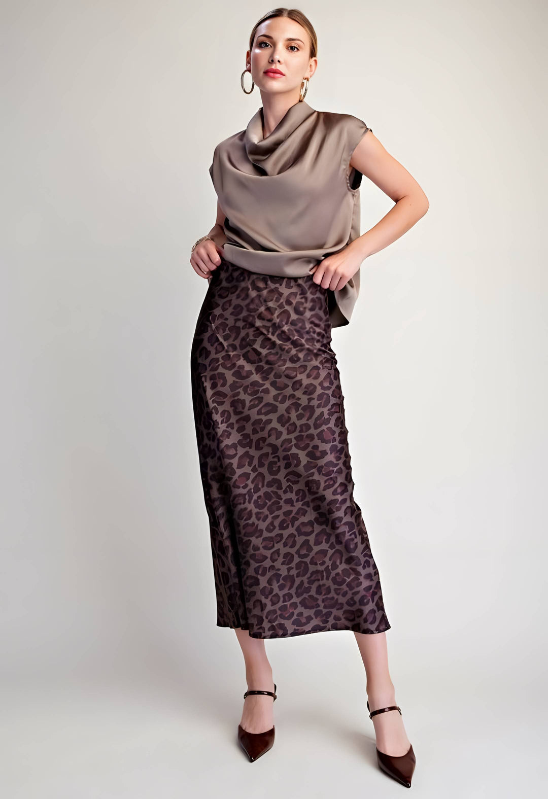 A model wearing a brown and black leopard-print midi skirt paired with a taupe cowl-neck top and dark brown pointed-toe heels, standing against a light gray studio background.
