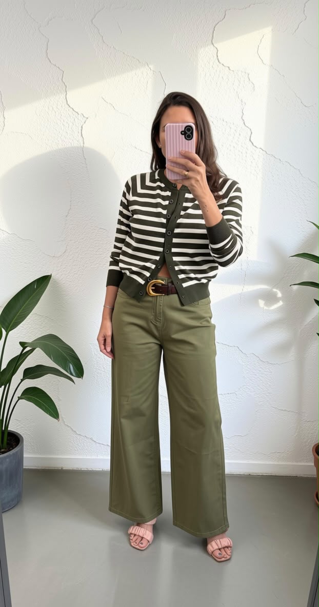 A woman takes a mirror selfie wearing a black and white striped cardigan, olive green wide-leg pants, a brown belt, and pink sandals, standing in a room with a white wall and potted plants.